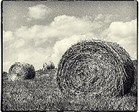 Three bales of hay sitting on a grassy hill with clouds in the sky