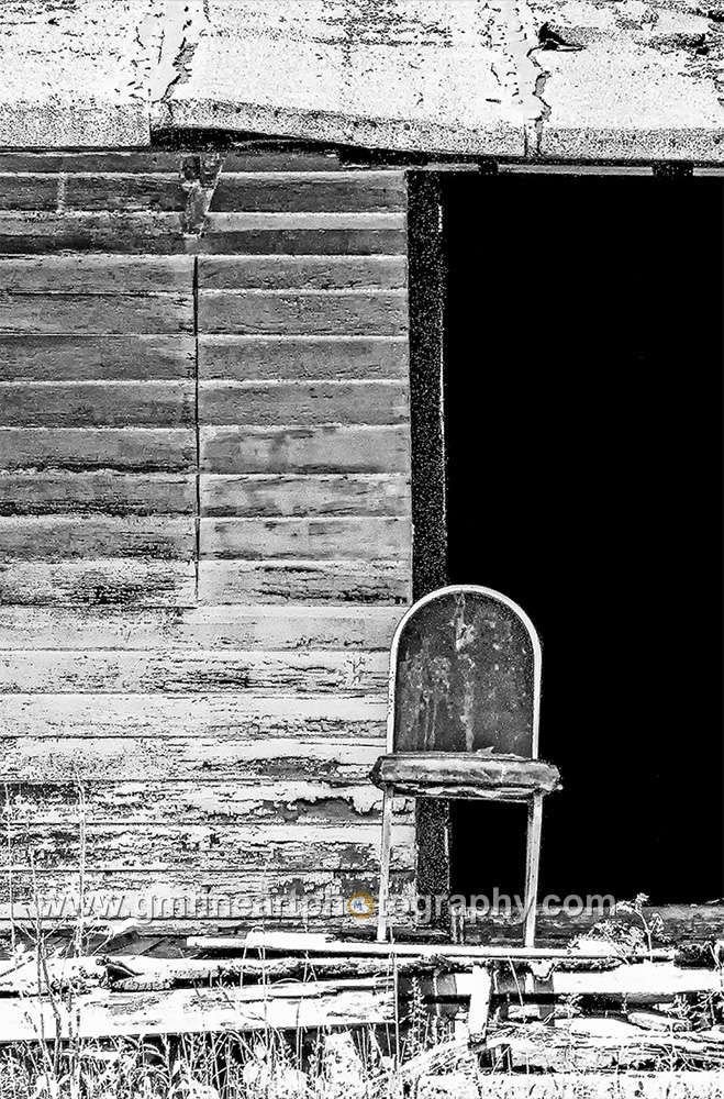 A lone old rickety chair in front of an old cabin with a black darkened doorway